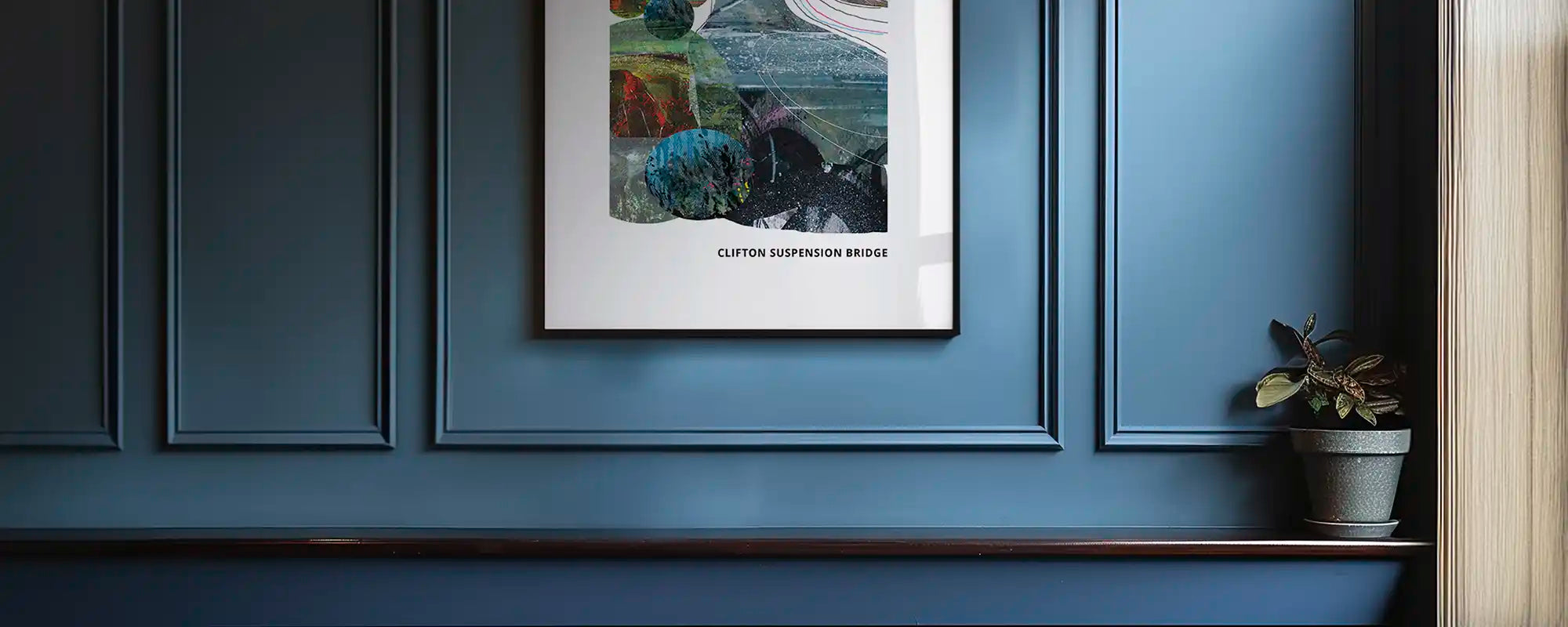 A framed art print titled 'Clifton Suspension Bridge' hangs on a dark blue panelled wall above a shelf with a small potted plant.