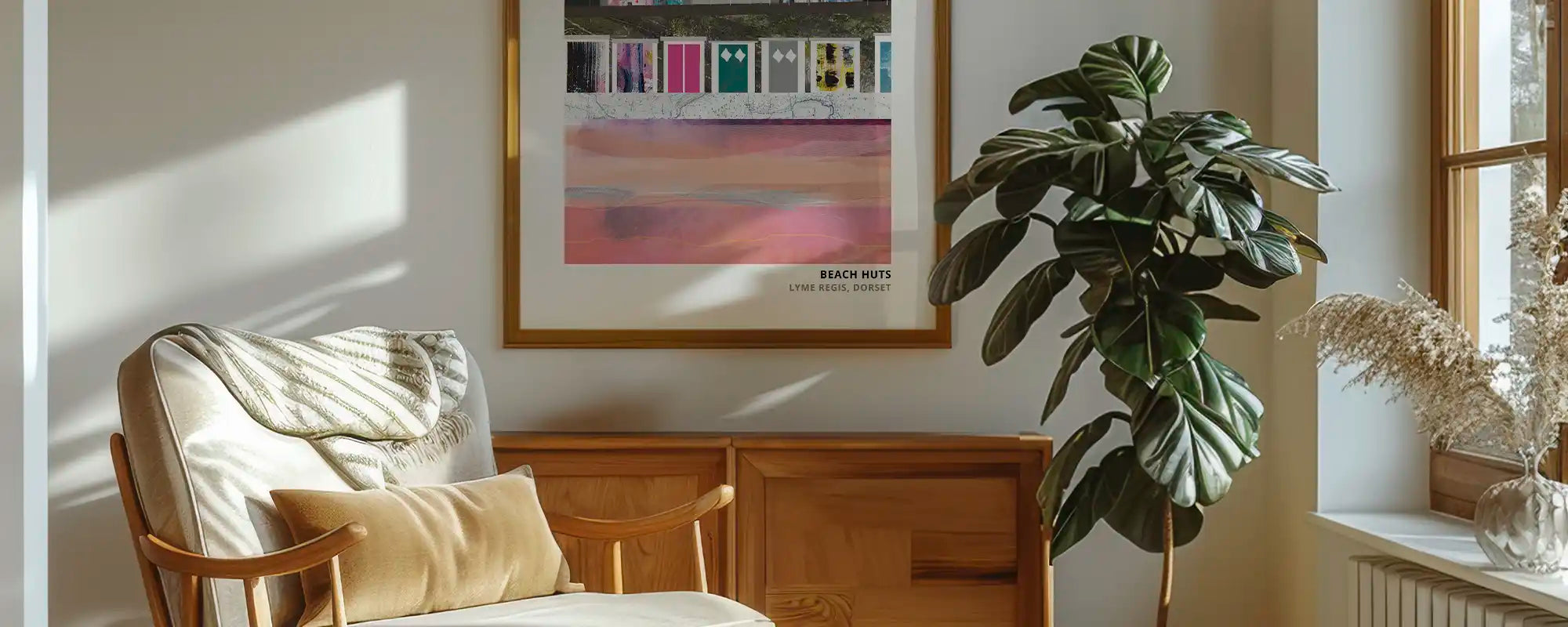 A framed art print featuring a colourful beach scene with the text 'BEACH WITH LOVE' and 'JERSEY', displayed on a beige wall in a sunlit room next to a wooden chair and a large green plant.