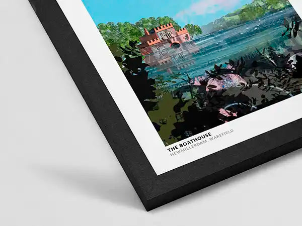 A framed art print of 'The Boathouse' by Richard Burns-Waitefield, featuring a colorful landscape illustration with a red boathouse, blue water, and green foliage, shown at an angle on a light gray background.