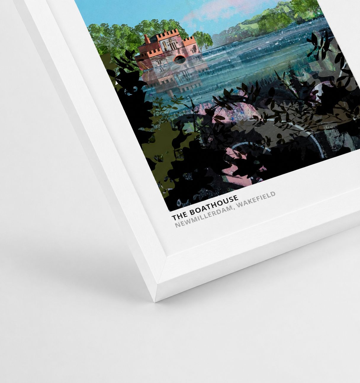 A framed art print of 'The Boathouse' in Newmillerdam, Wakefield, shown at an angle on a clean white background. The print features a colorful illustration of a red boathouse on blue water surrounded by green trees, enclosed in a thick white frame with black text at the bottom.
